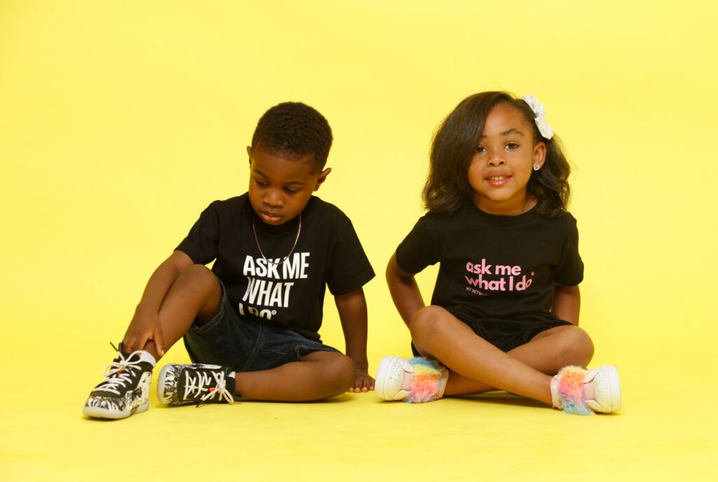 Deux enfants assis sur un fond jaune, portant des t-shirts noirs avec les textes « ASK ME WHAT I DO » et « ask me what I do », issus d'une collection en coton bio et matériaux recyclés.