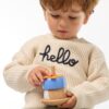Petit garçon portant un pull beige en coton bio avec l’inscription "hello", tenant un jouet en bois coloré fabriqué à partir de matériaux recyclés.