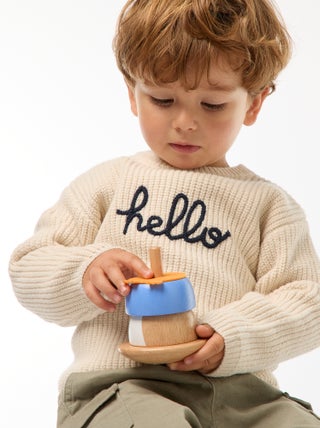 Petit garçon portant un pull beige en coton bio avec l’inscription "hello", tenant un jouet en bois coloré fabriqué à partir de matériaux recyclés.