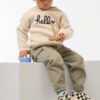 Petit garçon portant un pull beige en coton bio avec l’inscription "hello", tenant un jouet en bois coloré fabriqué à partir de matériaux recyclés.