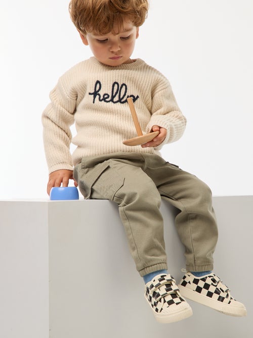 Petit garçon portant un pull beige en coton bio avec l’inscription "hello", tenant un jouet en bois coloré fabriqué à partir de matériaux recyclés.
