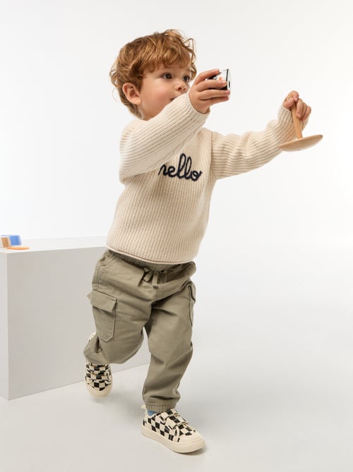 Petit garçon portant un pull beige en coton bio avec l’inscription "hello", tenant un jouet en bois coloré fabriqué à partir de matériaux recyclés.