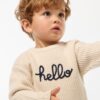 Petit garçon portant un pull beige en coton bio avec l’inscription "hello", tenant un jouet en bois coloré fabriqué à partir de matériaux recyclés.