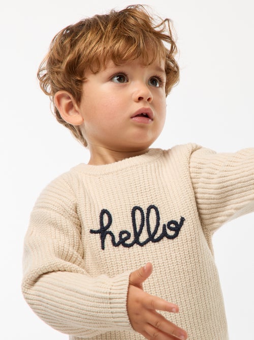 Petit garçon portant un pull beige en coton bio avec l’inscription "hello", tenant un jouet en bois coloré fabriqué à partir de matériaux recyclés.