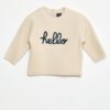 Petit garçon portant un pull beige en coton bio avec l’inscription "hello", tenant un jouet en bois coloré fabriqué à partir de matériaux recyclés.