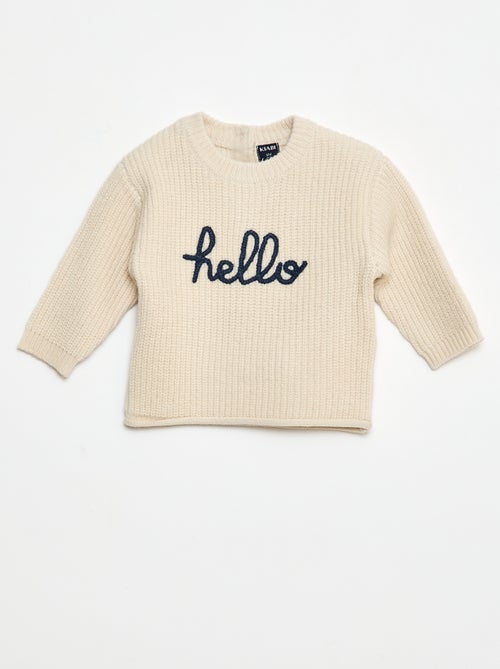 Petit garçon portant un pull beige en coton bio avec l’inscription "hello", tenant un jouet en bois coloré fabriqué à partir de matériaux recyclés.