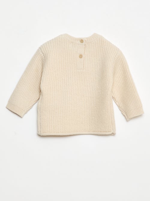 Petit garçon portant un pull beige en coton bio avec l’inscription "hello", tenant un jouet en bois coloré fabriqué à partir de matériaux recyclés.