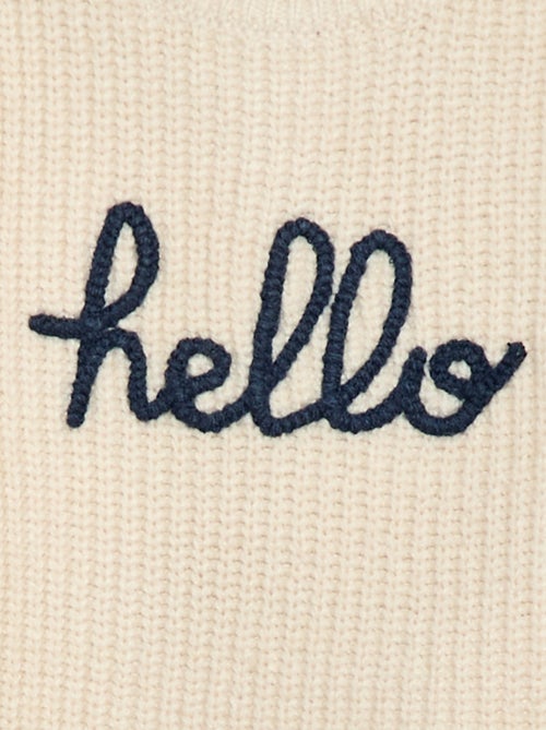 Petit garçon portant un pull beige en coton bio avec l’inscription "hello", tenant un jouet en bois coloré fabriqué à partir de matériaux recyclés.