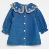 Robe-chemise enfant en denim bleu durable, avec boutons blancs et grand col rond brodé de motifs floraux, fabriquée en coton bio et matières recyclées.